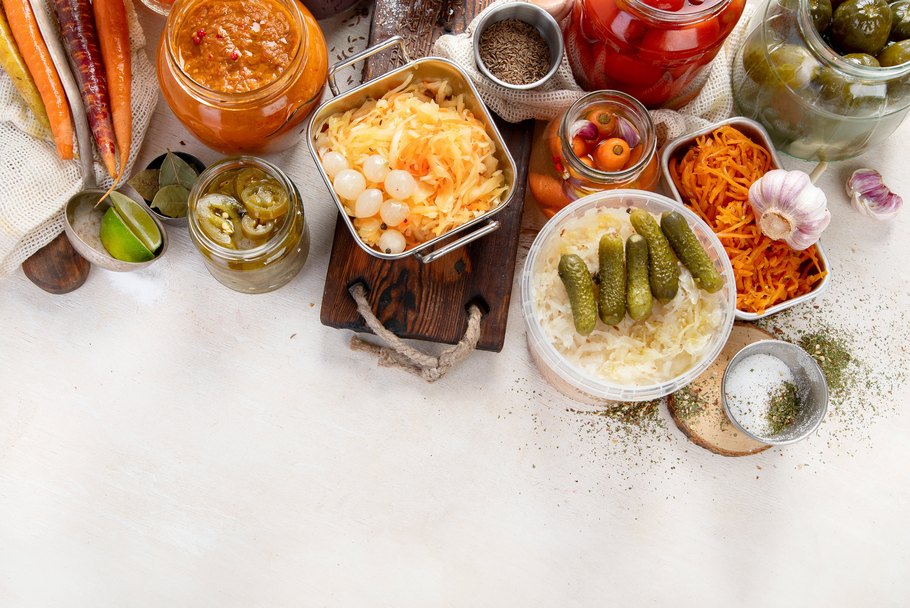 Fermentiertes Kraut in einem Behälter auf einem Holzbrett und verschiedene Arten von eingelegtem Gemüse in Gläsern.