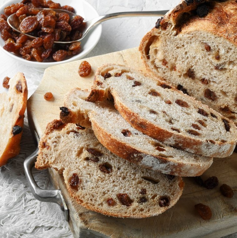 Rosinenbrot mit aufgeschnittenen Scheiben auf einem Brett auf weißer Unterlage, vor einem Schälchen mit Rosinen